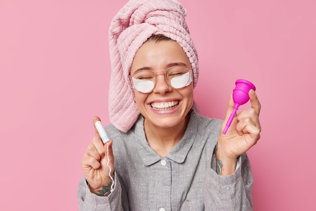 Positiva jovem europeia usa pijama confortável e toalha enrolada na cabeça segura tampão e copo de menstruação durante a menstruação isolada sobre fundo rosa aplica manchas de beleza sob os olhos