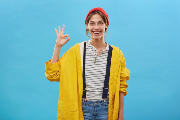 Positiva jovem fêmea vestida casualmente, mostrando o sinal de ok com a mão, aprovando algo. Mulher de casaco amarelo solto e chapéu vermelho isolado sobre a parede azul gesticulando com a mão. Trabalhadora alegre