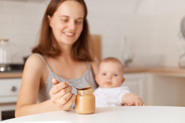 Foto grátis positiva sorridente jovem adulta de cabelos escuros mãe alimentando sua filha com purê de frutas ou vegetais, segurando a colher com alimentos saudáveis, posando na cozinha.