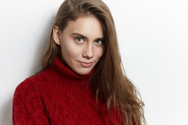 Positivo, alegre, atraente, mulher jovem, com cabelo solto, olhando com satisfação, sorrindo divertidamente, sendo feliz, vestida com um suéter de malha marsala de gola alta, posando para uma parede de stuido em branco