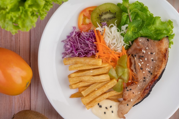 Posta de peixe com batatas fritas, kiwi, alface, cenoura, tomate e repolho em um prato branco.