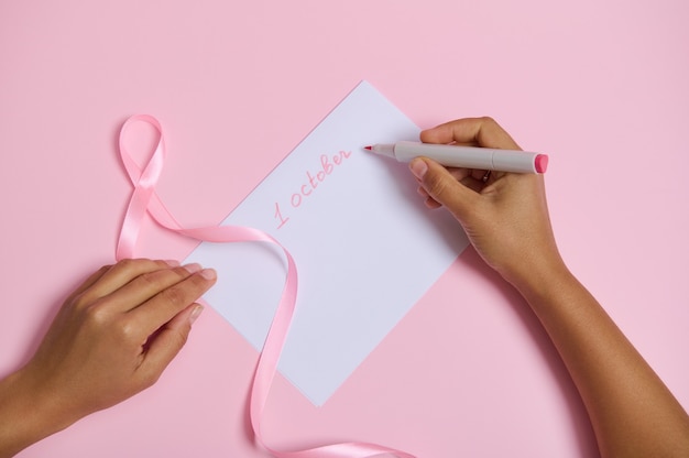 Postura plana da mão de uma mulher segurando uma caneta de feltro, escreve 1 de outubro em uma folha de papel em branco, símbolo de fita rosa do mês de outubro de conscientização sobre o câncer de mama, deitada sobre um fundo rosa com espaço para texto