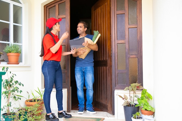 Postwoman latino segurando a área de transferência e entregando o pedido. Entregadora feliz em uniforme vermelho, falando com o cliente e entregando legumes em saco de papel. Serviço de entrega de comida e pós-conceito