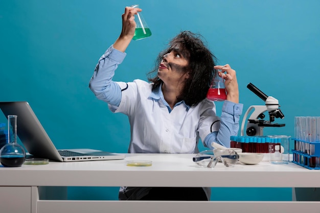 Potrait de químico bobo louco analisando frascos de vidro cheios de soros de experimentos líquidos enquanto está sentado na mesa. Cientista maníaco de aparência pateta tolo tendo copos cheios de compostos químicos.