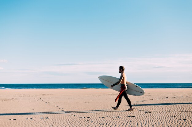 Praia aberta e vazia com surfista solitário em traje de mergulho, caminhar em direção à costa do mar ou oceano com prancha de surf