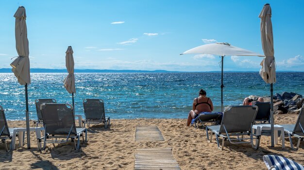 Praia na costa do mar Egeu com guarda-sóis e espreguiçadeiras, casal descansando e rochas perto da água em Nikiti, Grécia