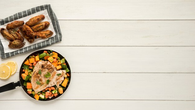 Prato com asas de frango e frigideira de legumes na mesa de madeira