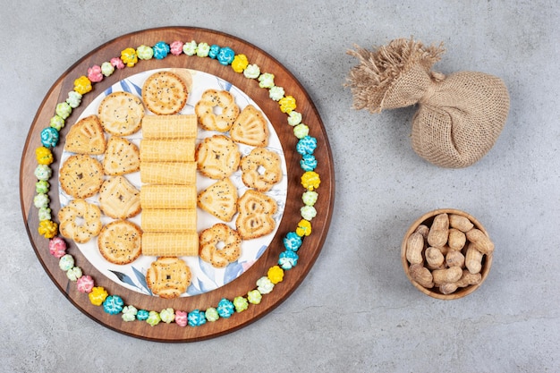 Prato de biscoitos variados rodeado por doces de pipoca na placa de madeira ao lado de um saco e uma tigela de amendoim na superfície de mármore.