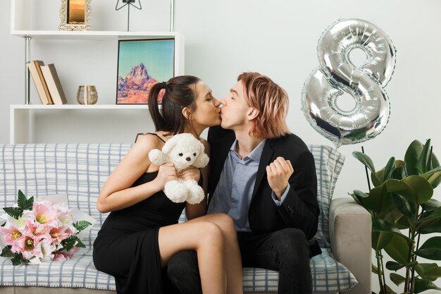 Prazer em beijar um ao outro jovem casal no feliz dia da mulher com o ursinho de pelúcia sentado no sofá da sala de estar