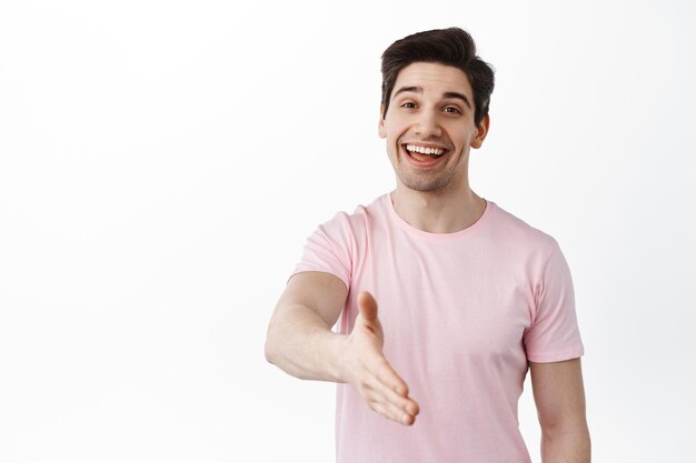 Prazer em conhecê-la. Homem caucasiano sorridente estende a mão para um aperto de mão, cumprimentando você, diga olá, se apresente com um sorriso amigável, em pé sobre um fundo branco