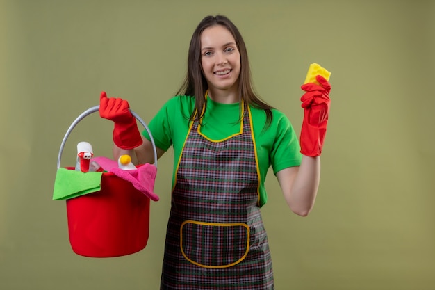 Foto grátis prazer em limpar jovem vestindo uniforme com luvas vermelhas segurando ferramentas de limpeza com uma esponja no fundo verde isolado