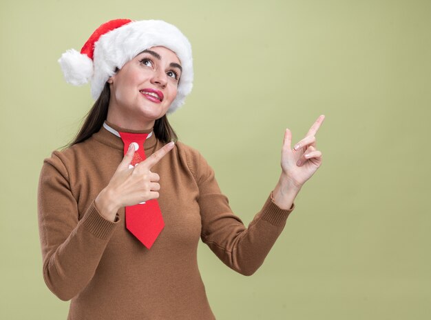 Prazer em olhar para o lado jovem linda com chapéu de natal e máscara médica apontando para o lado isolado em fundo verde oliva