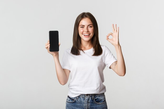 Prazer em sorrir menina morena mostrando a tela do smartphone e mostrando o gesto ok em aprovação.