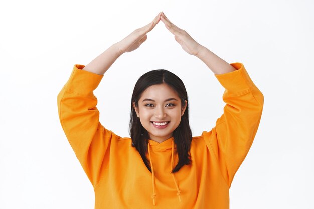 Preenchimento de garota garantido. Retrato de close-up fofa boba mulher asiática com capuz laranja fazendo telhado com as mãos acima da cabeça sorrindo alegremente, conceito de segurança