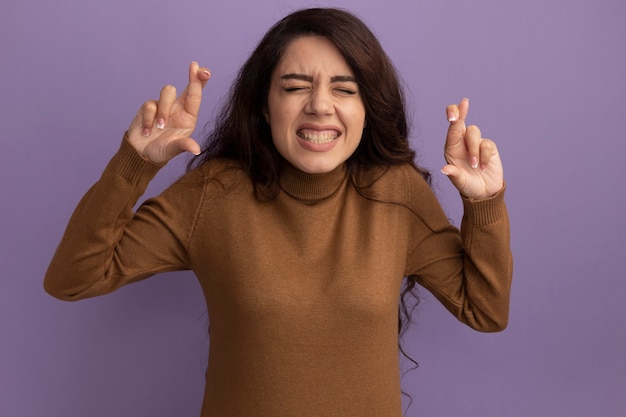 Preocupada com os olhos fechados, bela jovem vestindo uma blusa de gola alta marrom cruzando os dedos isolados na parede roxa