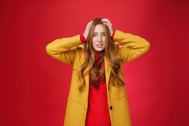 Preocupada e assustada, garota ruiva segurando as mãos na cabeça, fazendo caretas preocupada e preocupada, cerrando os dentes, franzindo a testa preocupada e se sentindo nervosa pelo projeto que vai falhar sobre o fundo vermelho.