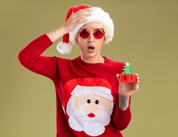 preocupada jovem loira usando chapéu de Natal e suéter de Natal de Papai Noel com óculos segurando o brinquedo da árvore de Natal com data procurando manter a mão na cabeça isolada na parede verde oliva