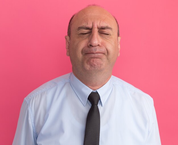 Preocupado com os olhos fechados, homem de meia-idade vestindo camiseta branca com gravata isolada na parede rosa
