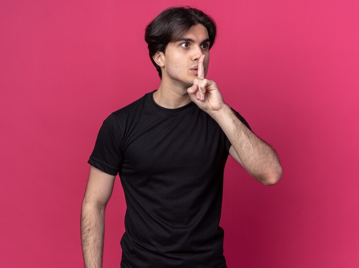Foto grátis preocupado em olhar para o lado jovem bonito vestindo camiseta preta mostrando gesto de silêncio isolado na parede rosa
