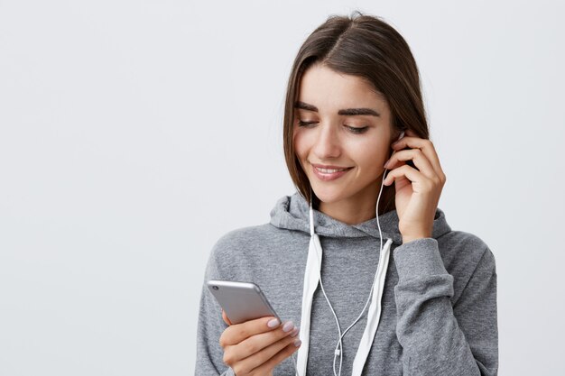 Preparando-se para a corrida matinal. Estilo de vida urbano. Retrato de uma jovem bonita caucasiana com cabelos escuros no capuz esportivo cinza, usando fones de ouvido, pesquisando a lista de reprodução favorita no smartphone.