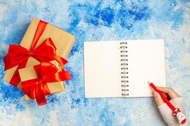 Presente de Natal de vista superior amarrado com caneta de Papai Noel de bloco de notas de laço vermelho na mão feminina na mesa azul