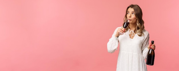 Primavera de férias e conceito de festa Retrato de mulher loira bonita independente despreocupada bebendo vinho de vidro segurando garrafa e degustação de bebida olha para cima comemorando com amigos fundo rosa