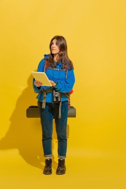 Procurando o caminho com o mapa. Retrato de uma garota alegre jovem turista caucasiano com bolsa e binóculos isolados no fundo amarelo do estúdio. Preparando-se para viajar. Resort, emoções humanas, férias.