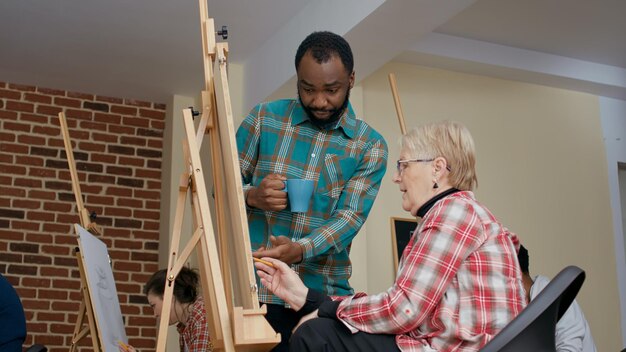 Professor afro-americano orientando mulher sênior para desenhar esboço na tela. Jovem artista ensinando nova técnica de desenho ao aluno mais velho para criar design de obra-prima na aula de arte.