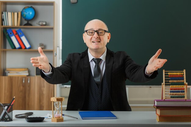 Foto grátis professor de homem usando óculos com registro de classe sentado na mesa da escola na frente do quadro-negro na sala de aula olhando para a câmera feliz e satisfeito abrindo as mãos gesto de boas-vindas