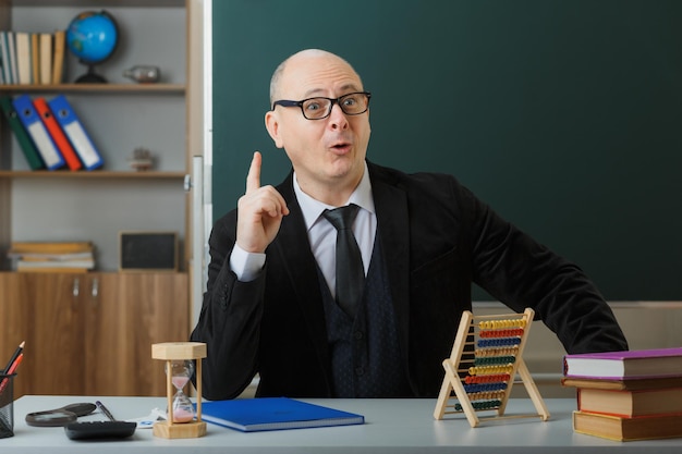 Professor de homem usando óculos com registro de classe sentado na mesa da escola na frente do quadro-negro na sala de aula usando ábaco parecendo surpreso apontando com o dedo indicador para cima tendo uma ótima ideia