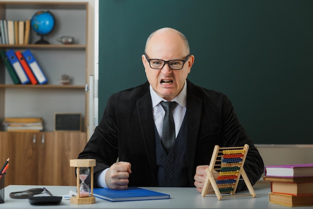 Professor de homem usando óculos com registro de classe sentado na mesa da escola na frente do quadro-negro na sala de aula usando os punhos cerrados do ábaco gritando com expressão agressiva