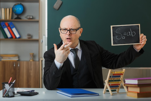 Professor de homem usando óculos sentado na mesa da escola na frente do quadro-negro na sala de aula mostrando a lousa explicando a lição sendo descontente e com raiva