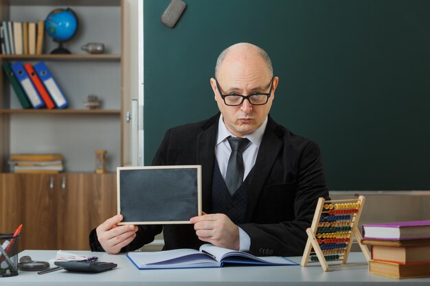 Professor de homem usando óculos sentado na mesa da escola na frente do quadro-negro na sala de aula mostrando lousa explicando a lição explicando a lição com cara séria