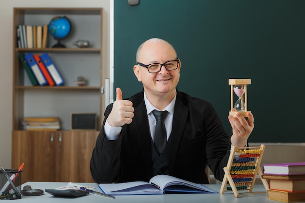 Foto grátis professor de homem usando óculos sentado na mesa da escola na frente do quadro-negro na sala de aula segurando a ampulheta explicando a lição mostrando o polegar feliz e satisfeito