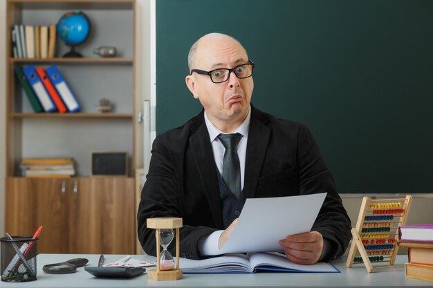 Professor de homem usando óculos sentado na mesa da escola na frente do quadro-negro na sala de aula verificando a lição de casa dos alunos olhando espantados e surpresos