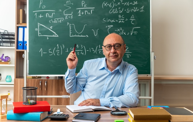 Professor de meia-idade impressionado, usando óculos, sentado à mesa com o material escolar segurando uma caneta na sala de aula