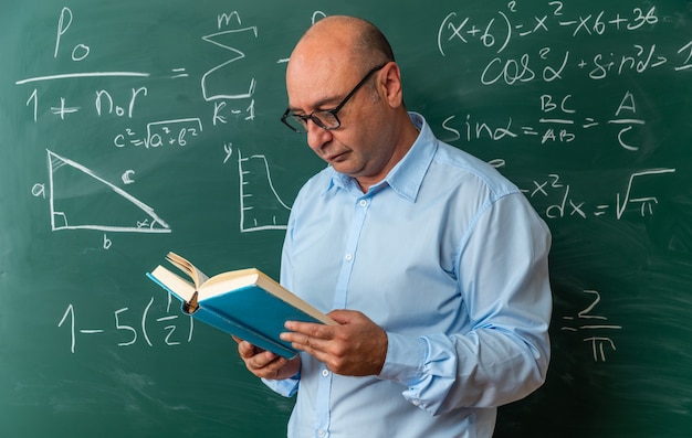 Professor de meia-idade pensando em usar óculos em pé na frente da lousa lendo um livro