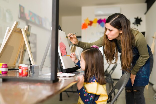 Professor hispânico atraente está ensinando uma menina bonita como usar o pincel em uma tela durante uma aula de arte