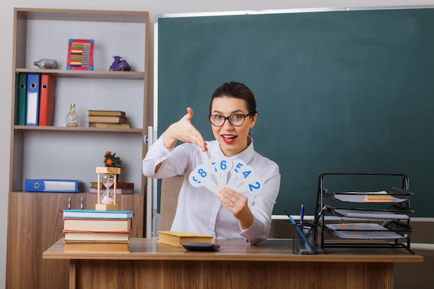 Professora jovem de óculos apresentando placas de matrícula explicando a lição feliz e positiva sorrindo sentado na mesa da escola na frente do quadro-negro na sala de aula