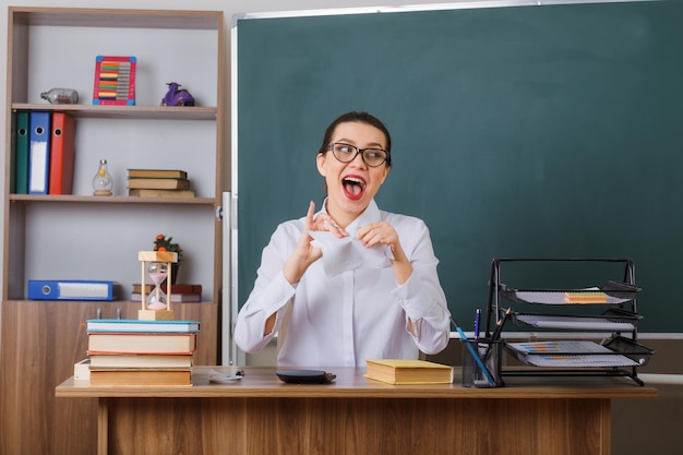 Professora jovem de óculos rasgando um pedaço de papel parecendo alegre sentado na mesa da escola na frente do quadro-negro na sala de aula