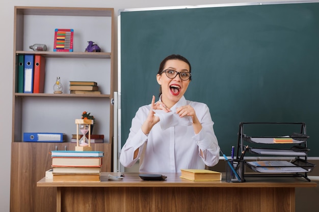 Professora jovem de óculos rasgando um pedaço de papel parecendo loucamente feliz sentado na mesa da escola na frente do quadro-negro na sala de aula