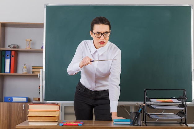 Professora jovem de óculos segurando o ponteiro enquanto explica a lição franzindo a testa em pé na mesa da escola na frente do quadro-negro na sala de aula