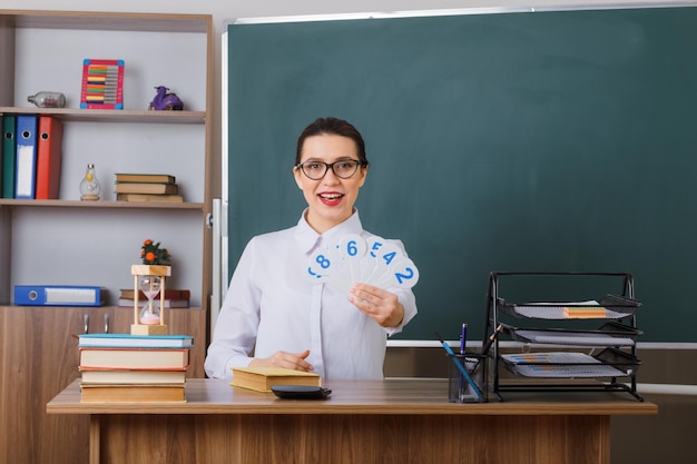 Professora jovem de óculos segurando placas explicando a lição sorrindo confiante sentado na mesa da escola na frente do quadro-negro na sala de aula