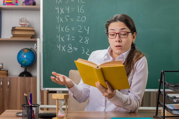 Professora jovem de óculos segurando um livro, preparando-se para a aula de leitura, sendo confusa sentada na mesa da escola em frente ao quadro-negro na sala de aula