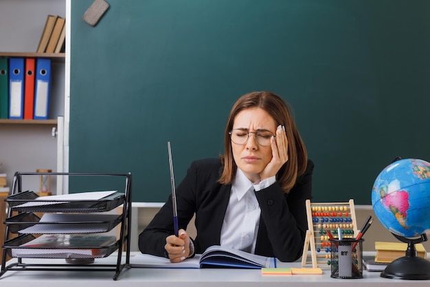 Professora jovem de óculos sentado na mesa da escola na frente do quadro-negro na sala de aula com ábaco e globo verificando o registro de classe segurando o ponteiro parecendo indisposto sentindo dor