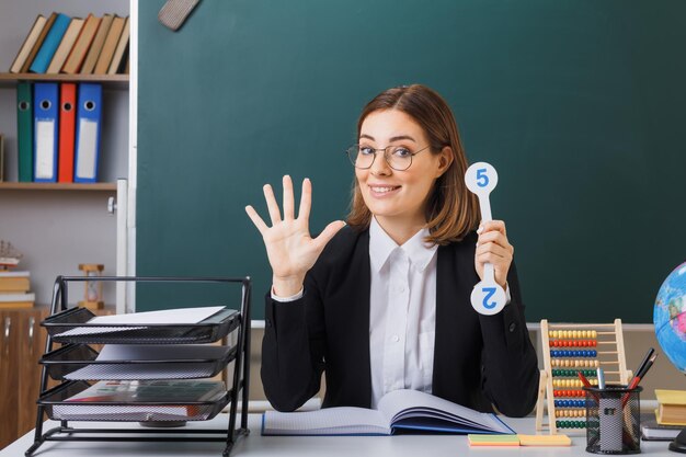 Professora jovem de óculos sentado na mesa da escola na frente do quadro-negro na sala de aula segurando placas explicando lição feliz e positiva mostrando o número cinco com a palma da mão aberta