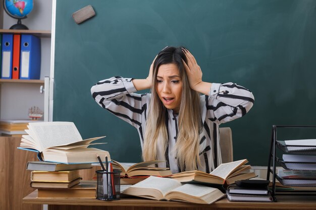 professora jovem sentada na mesa da escola na frente do quadro-negro na sala de aula com livros olhando bagunça na mesa dela sendo confusa e preocupada com as mãos na cabeça
