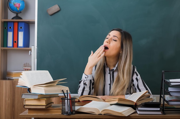 Professora jovem sentada na mesa da escola na frente do quadro-negro na sala de aula entre livros na mesa dela olhando cansado e sobrecarregado bocejando cobrindo a boca com a mão