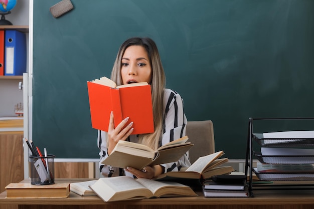 professora jovem sentada na mesa da escola na frente do quadro-negro na sala de aula segurando livros lendo olhando espantado e surpreso