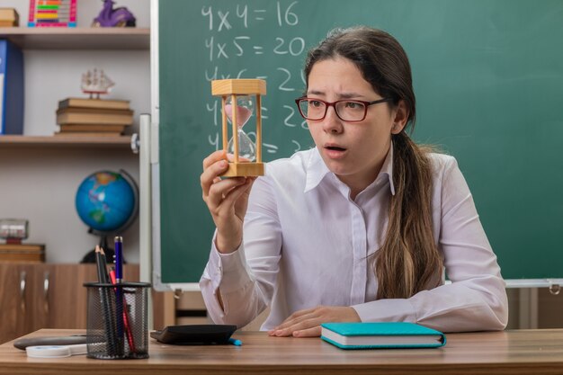 Professora jovem usando óculos com ampulheta olhando para ela surpresa se preparando para a aula sentada na mesa da escola em frente ao quadro-negro na sala de aula